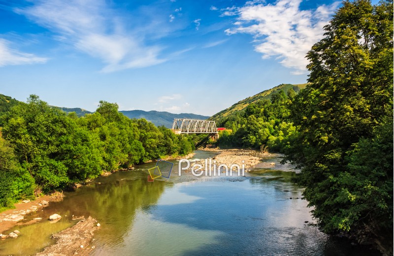 River flows among a green forest at the foot of the mountain. picturesque nature of rural area in Carpathians on a serene summer day. White metal train bridge in the distance. 