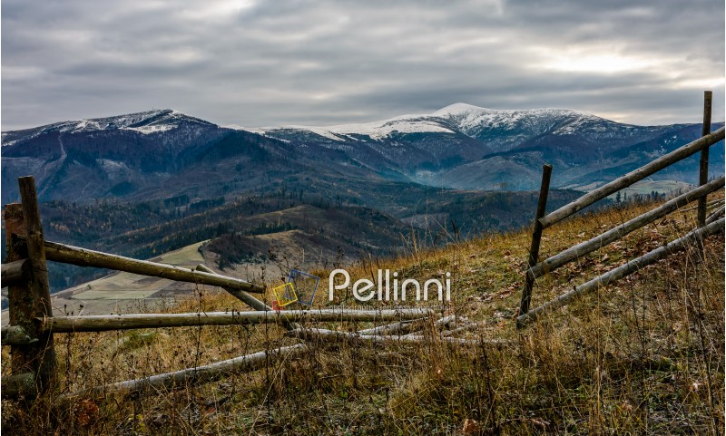 wooden fence on the hill with weathered grass. mountains with snowy peaks in the distance. Late autumn landscape in cloudy weather.
