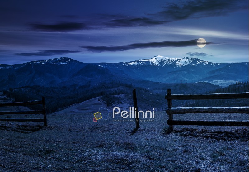wooden fence on grassy hillside near mountains with snowy peaks in spring at night in full moon light