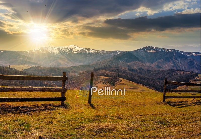 wooden fence on grassy hillside near mountains with snowy peaks in spring evening light