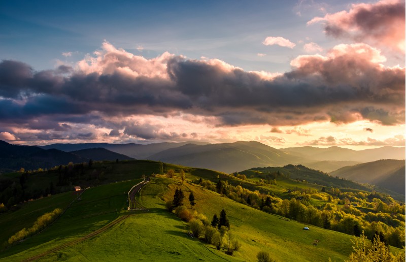 glorious cloudy sunset over rural area. beautiful springtime landscape with mountain ridge in the distance. country road winds through grassy slopes on rolling hill