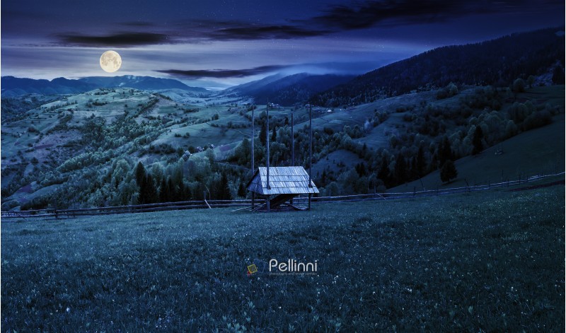 hay shed on a grassy field in mountains. beautiful countryside landscape in springtime at night in full moon light. village on the distant hills