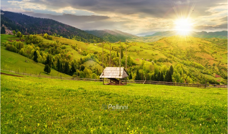 hay shed on a grassy field in mountains. beautiful countryside landscape in springtime at sunset in evening light. cloudy afternoon. village on the distant hills