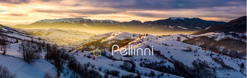 gorgeous panorama of mountainous countryside at sunrise in winter. snow covered rural area on rolling hills glow in morning sunlight. huge mountain ridge in a distance under cloudy sky