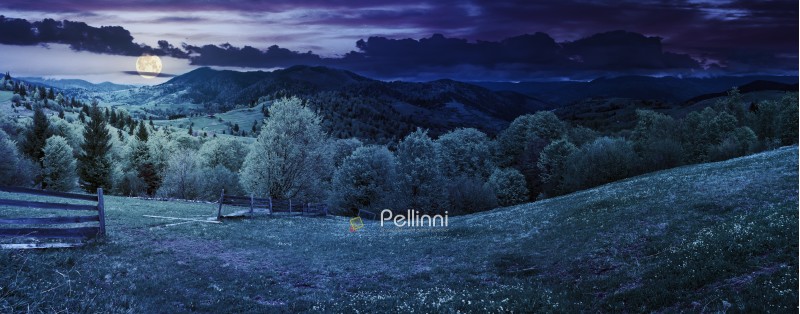 panorama of mountainous countryside in springtime at night in full moon light. beautiful highland landscape. wooden fence on the grassy field. row of trees along the hill. rural area in the distance