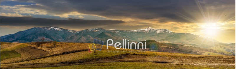 Early spring highland landscape. Panorama of rural fields on hill side in mountains with snowy peaks in evening light