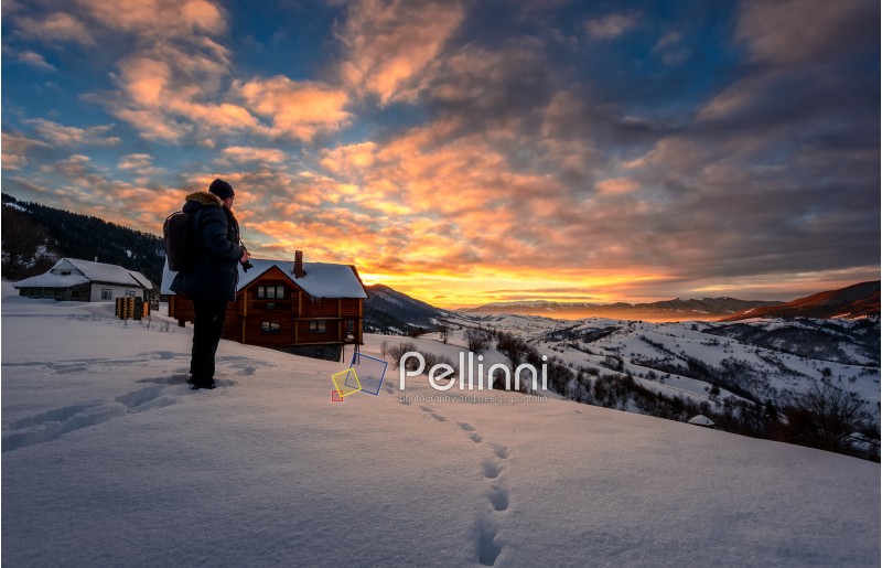 landscape photographer stands on the edge of hillside covered with snow near the village houses in mountainous rural area and looks on winter sunrise