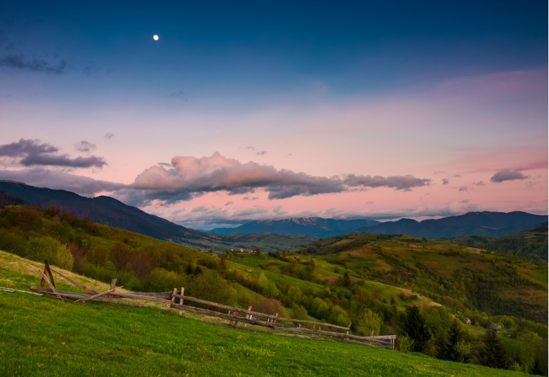 rural area at dusk with moon on cloudy sky. beautiful mountainous landscape with agricultural fields and wooden fences on grassy slopes in springtime