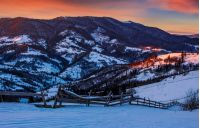 epic red sunrise in Carpathian countryside landscape. wooden fence through hillside with forest. Magical frosty morning.