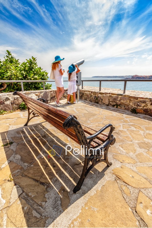 Mother and daughter standing on a seaside observation deck with a bench and looking into bincular