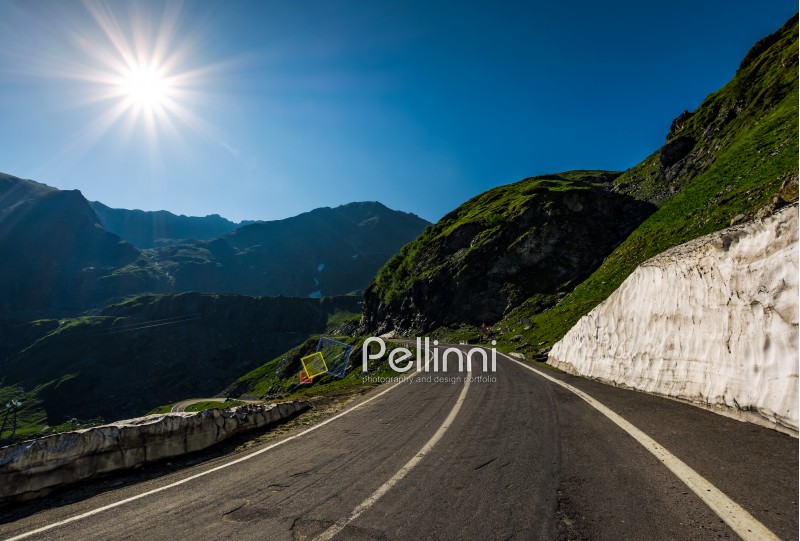 asphalt road uphill through mountain range. snow and grass on hillside under the clear blue sky with sun