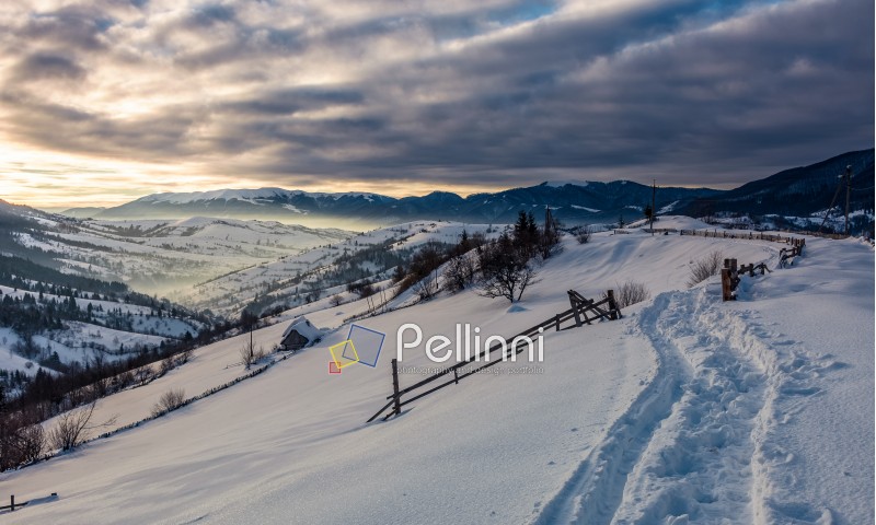 beautiful countryside in mountains. path in a snow near the wooden fence over the village and rural fields on hillsides covered with snow at sunrise