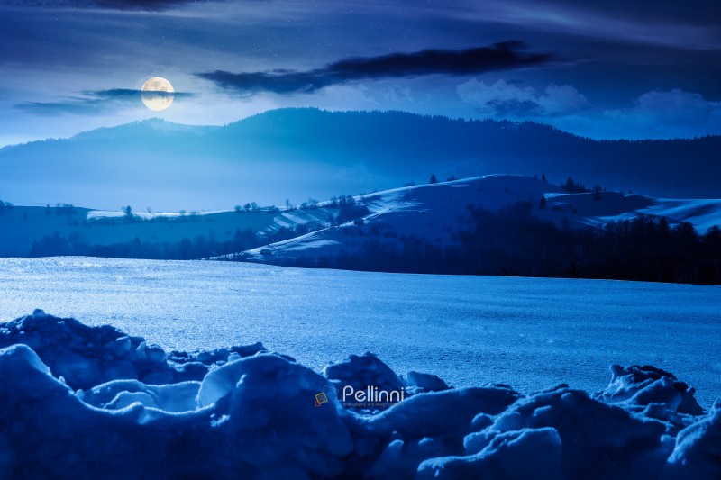beautiful winter landscape with snow covered rolling hills under dark sky at night. majestic mountains in full moon light. frosty countryside scenery. backdrop for fiction art or mystery concepts