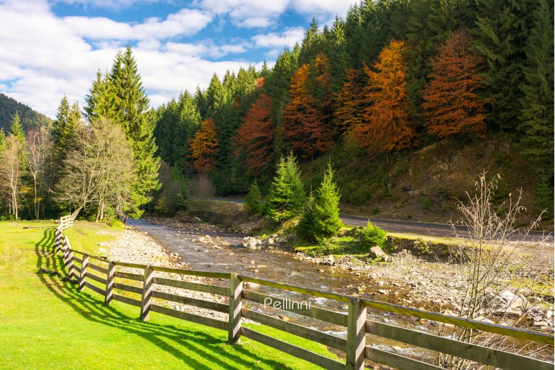 camping place near the river. wooden fence on a grassy lawn along the shore. road under the hill with trees in fall color. beautiful sunny autumn weather