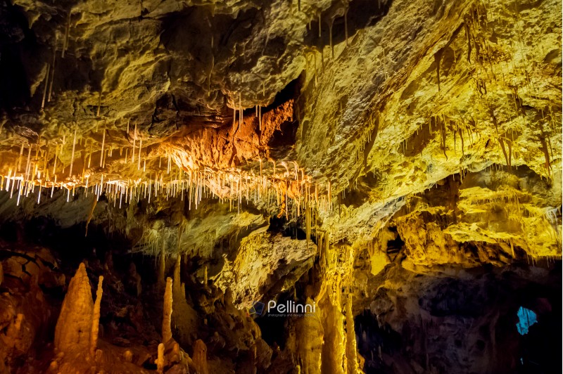 ceiling of cave with colorful textured walls and stalactites and stalagmite lit from behind