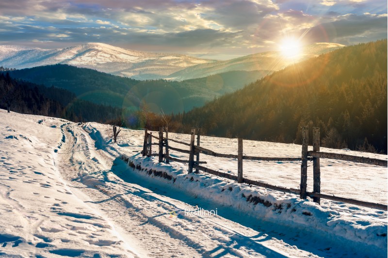 country road in to the winter mountains at sunset. wooden fence along the road. composite image