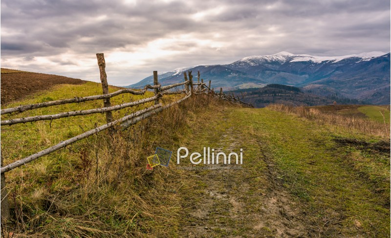 fence along dirt road in mountainous rural area. agricultural fields on hills in late autumn. mountain ridge with snowy tops in the distance
