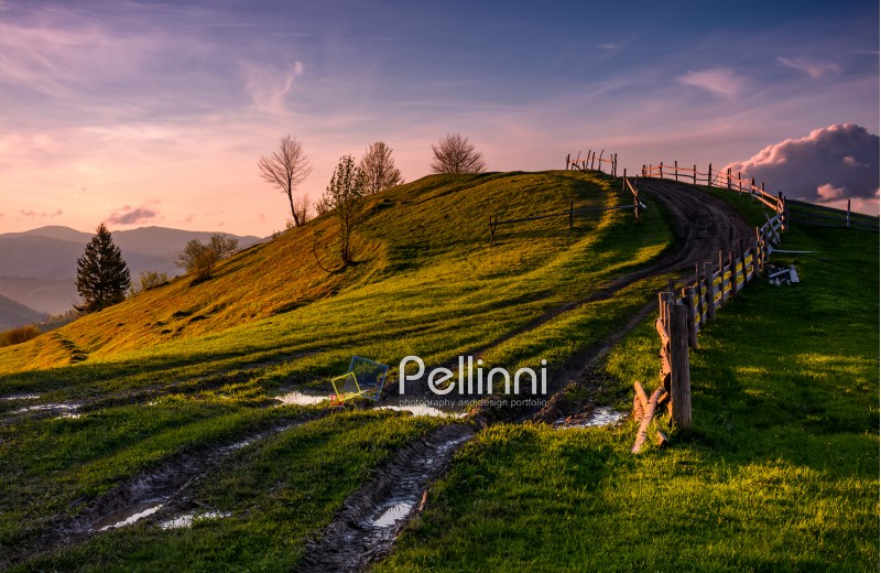 wooden fence along the country dirt road uphill the grassy knoll in springtime at dusk. Spectacular nature scenery in mountainous rural area with gorgeous pink sky with some clouds