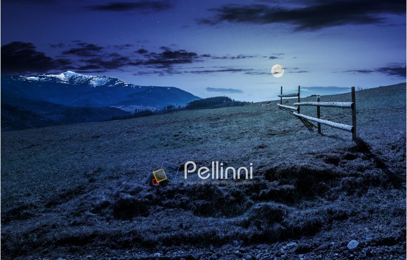wooden fence on grassy hillside near mountains with snowy peaks in spring at night in full moon light