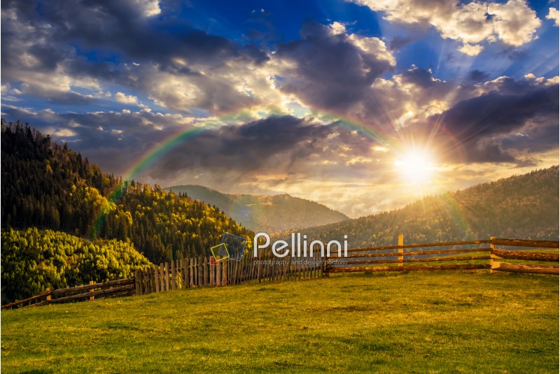 wooden fence through the grassy meadow in mountains. beautiful Carpathian countryside landscape with cloudy sky at sunset with rainbow