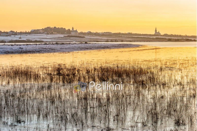 frozen river near village with snow on the shore and few churches in evening light