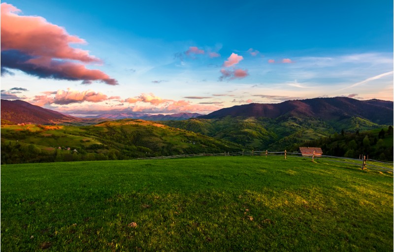 gorgeous mountainous countryside at dusk. beautiful landscape with rural fields, fence and  woodshed. pink clouds on a spring blue sky over the mountain ridge in the distance
