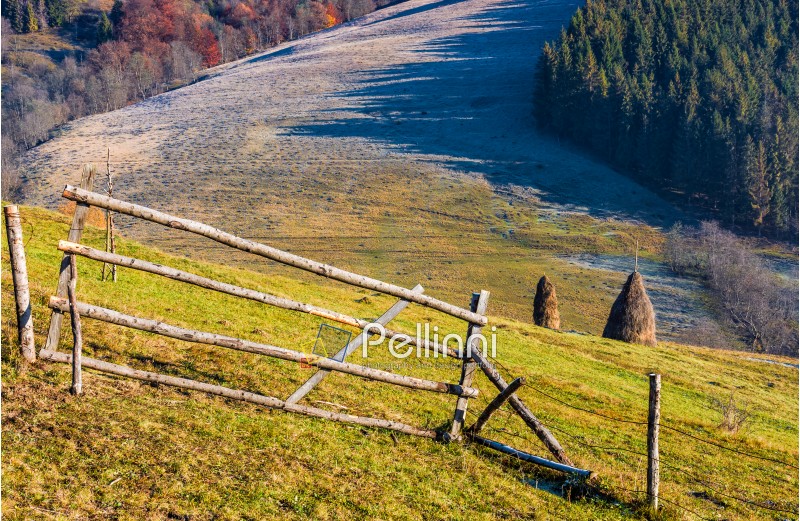haystack behind the fence on the rural field on hillside. frosty grass in forest shade at sunrise in autumn