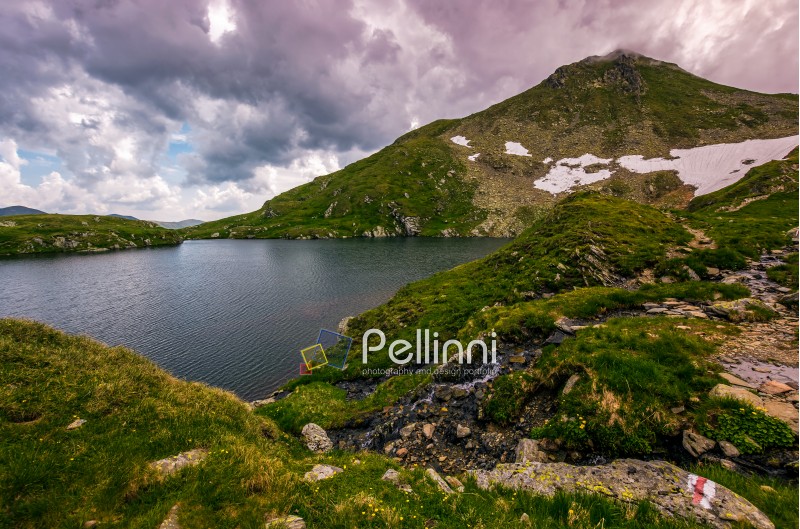 clear lake in mountains with snow and grass on rocky hillside. dramatic weather in picturesque summer scenery