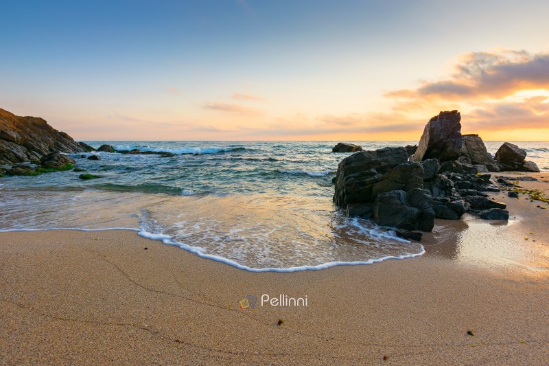 landscape by the sea at sunrise in summer. beautiful beach with rocks washed by calm waves in orange light. view to horizon under blue sky with clouds. scenic travel destination for leisure and relax