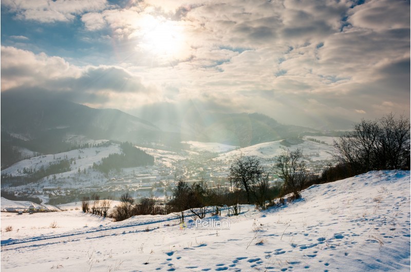 magical winter countryside. sun ray through the cloudy sky. snowy hill and leafless trees. village down in the valley