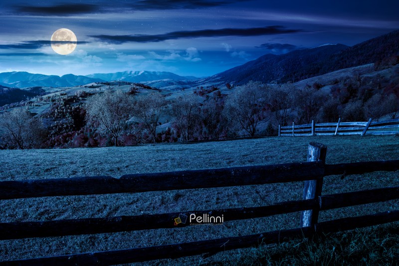 mountainous rural landscape in autumn at night. field behind the wooden fence in full moon light. scenery in fall season. forest on the hill. ridge in the distance