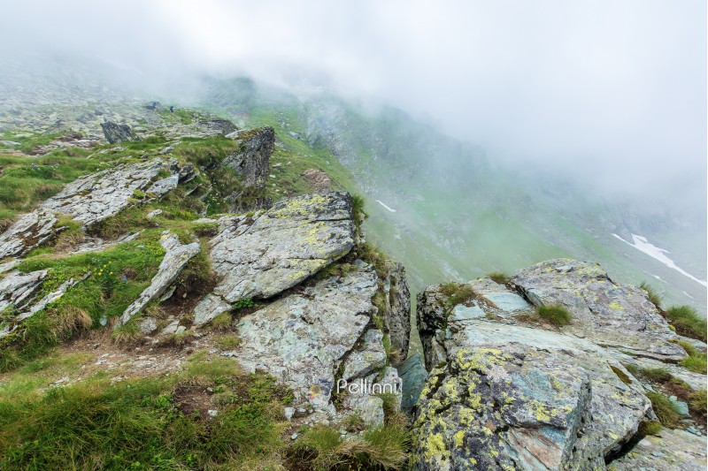 on the edge of rocky cliff of Fagaras ridge. mysterious scenery of Romanian highlands in thick fog. beautiful summer landscape. popular travel destination. couple of tourists in the distance