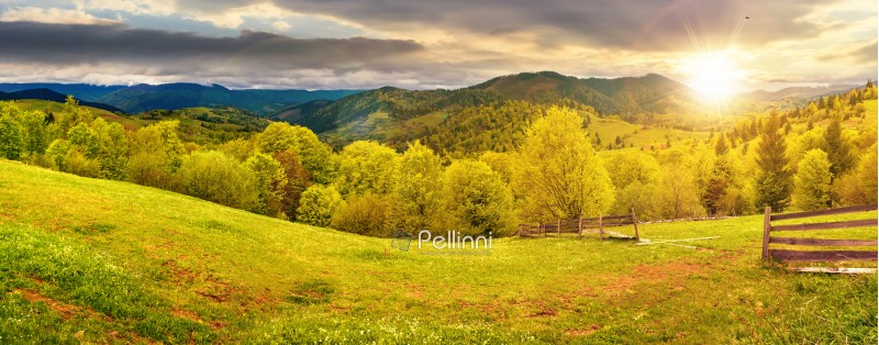 panorama of mountainous countryside in springtime at sunset. beautiful highland landscape. wooden fence on the grassy field. row of trees along the hill. rural area in the distance. cloudy sky