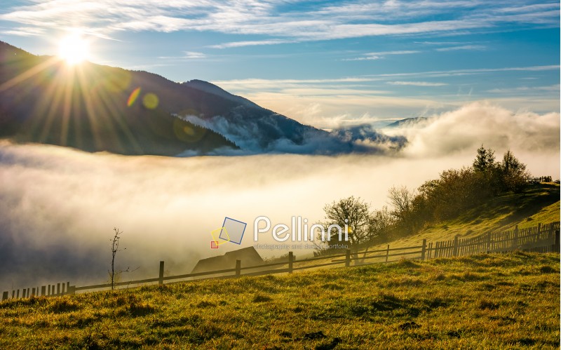 rural valley at foggy sunrise in autumn. wooden fence on the hillside with woodshed and orchard. gorgeous autumnal landscape with high mountains in a distance.