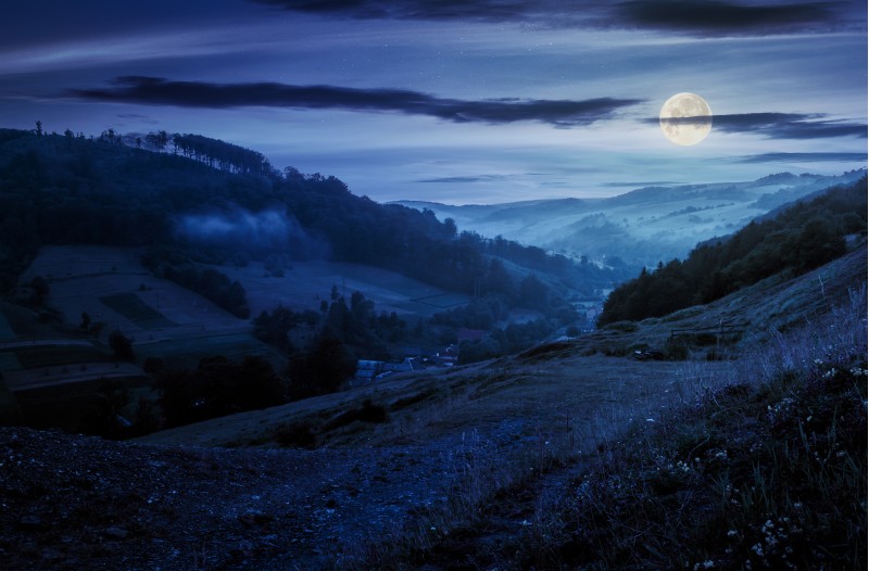 rural valley with forested hills at night in full moon light. beautiful summer landscape in Carpathian mountains