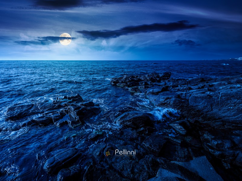sea coast with stones and boulders at night. view of beach with sea coast with stones and boulders at night. view of beach with rock in seaweed. full moon over dynamic cloudscape on the sky above horizon. mysterious summer vacations background concept