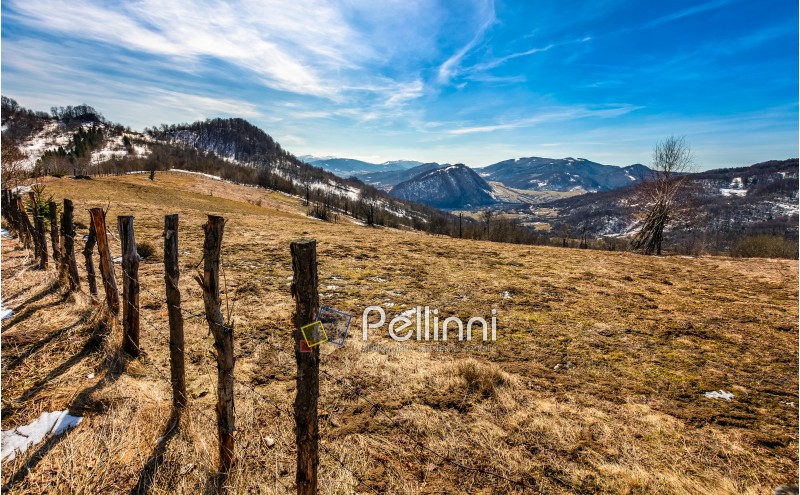 spring has sprung in rural area. wooden fence on agricultural field, yellow weathered grass covered with snow. village in the distance at the mountain ridge foot. nature on sunny day under blue sky