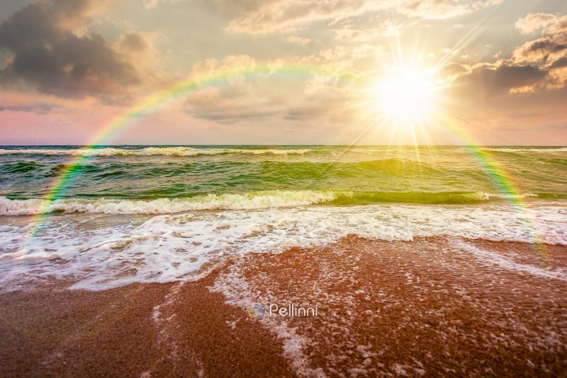 stormy weather on the sandy beach at sunset. dramatic sky over the sea in evening light. moody summer landscape. green waves crashing the shore. majestic cloudscape and rainbow above the horizon