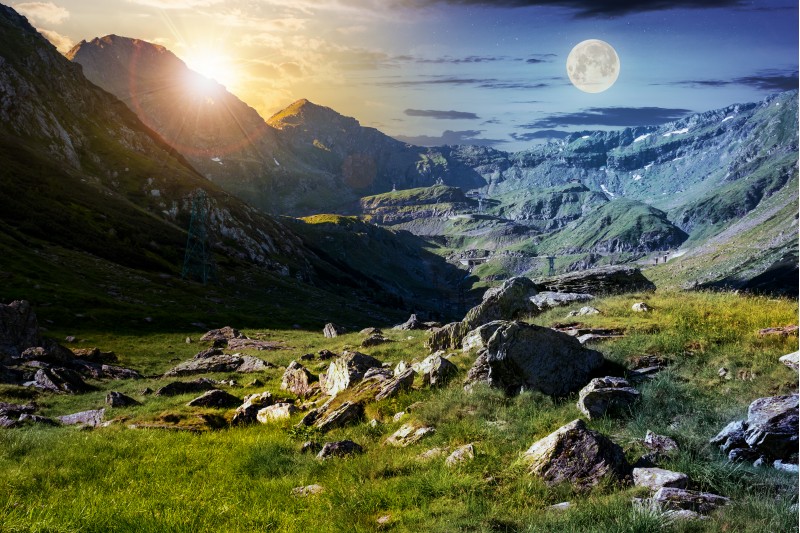 time change concept in Transfagarasan valley. rocks on grassy meadow and slopes lit by sun and moon simultaneously. half of the valley in shade of mountain ridge