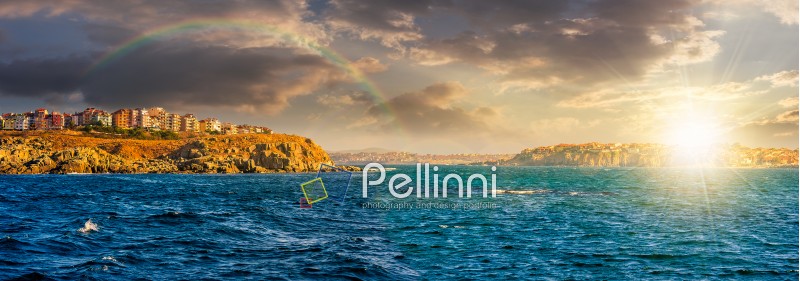 composite summer seascape. panoramic view of old resort town under the rainbow on a rocky cliff above the seashore. blue and calm water in the sea at sunset;