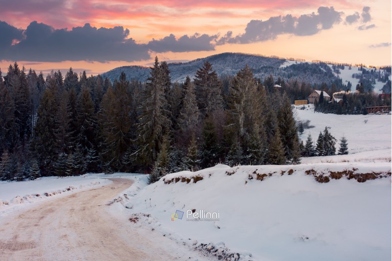 wonderful winter countryside in mountains at dusk. road winds down the slope in to the forest. village on a snowy hill in the distance. beautiful evening sky with clouds