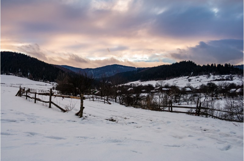 wooden fence along the snowy road. beautiful winter landscape of mountainous rural area at cloudy sunset
