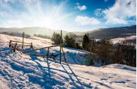 broken fence on a snowy slope in rural are. lovely winter landscape with bright sun over the mountain ridge