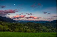 countryside landscape in mountains at dusk with moonrise. grassy meadow on a hillside behind the fence. spectacular evening sky with red clouds and moon