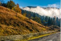 countryside road and fence at the mountain foot. cloud rising up from the forest at autumn morning