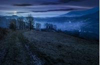 dirt road to village down the hill. trees on hillside and village in valley in autumnal countryside landscape at night in full moon light