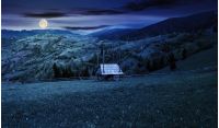 hay shed on a grassy field in mountains. beautiful countryside landscape in springtime at night in full moon light. village on the distant hills