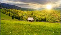hay shed on a grassy field in mountains. beautiful countryside landscape in springtime at sunset in evening light. cloudy afternoon. village on the distant hills