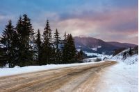 Empty asphalt mountain road under the snow  near the coniferous forest with cloudy sky in winter evening light