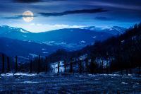 mountainous countryside at night. winter to spring season change scenery. melting snow and leafless trees on the rolling hills in full moon light. view to the distant valley